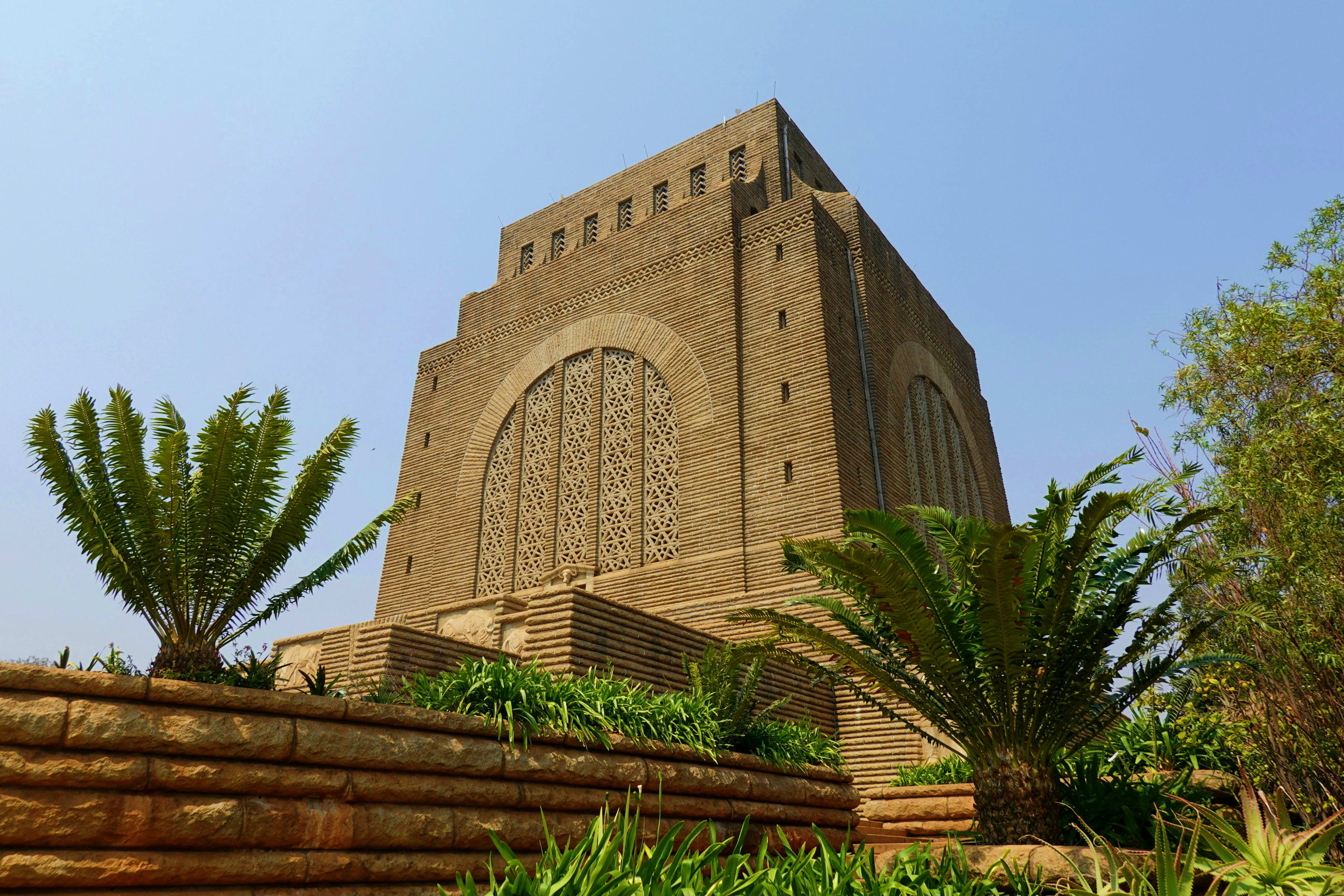 South Africa Voortrekker Monument brick tower with ornate geometric patterns and decorative windows, surrounded by palm trees and garden w